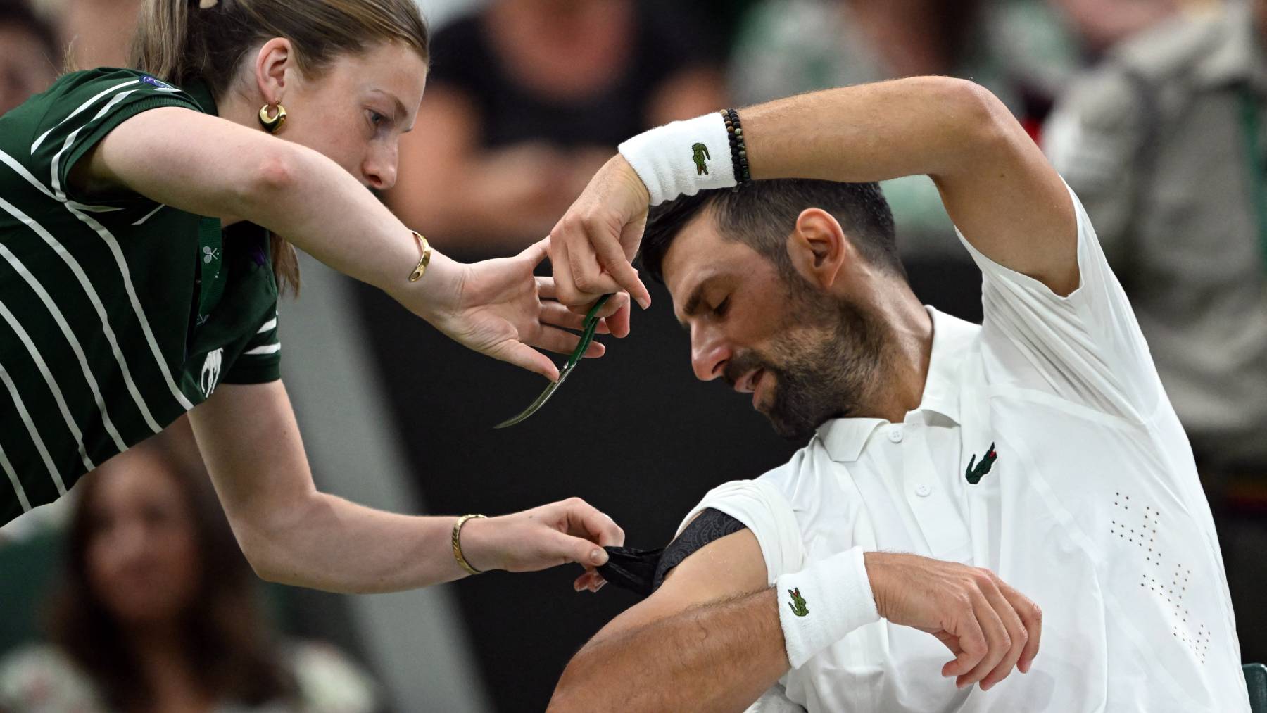 A member of the ground staff helps Serbia's Novak Djokovic to cut a piece of his kinesio tape during a break of his men's singles second round tennis match against Greece's Stefanos Tsitsipas on the third day of the 2026 Wimbledon Championships at The All England Lawn Tennis and Croquet Club in Wimbledon, southwest London, on July 1, 2026.