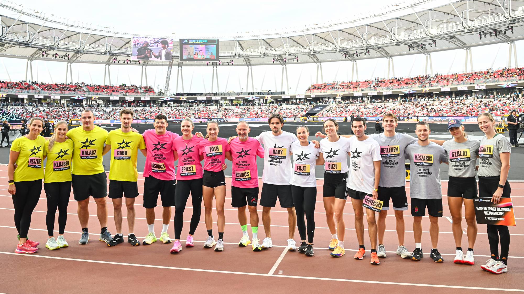 Group of runners in bright yellow, pink, white, and gray shirts pose arm-in-arm on a stadium track, with a large crowd in the stands behind them.